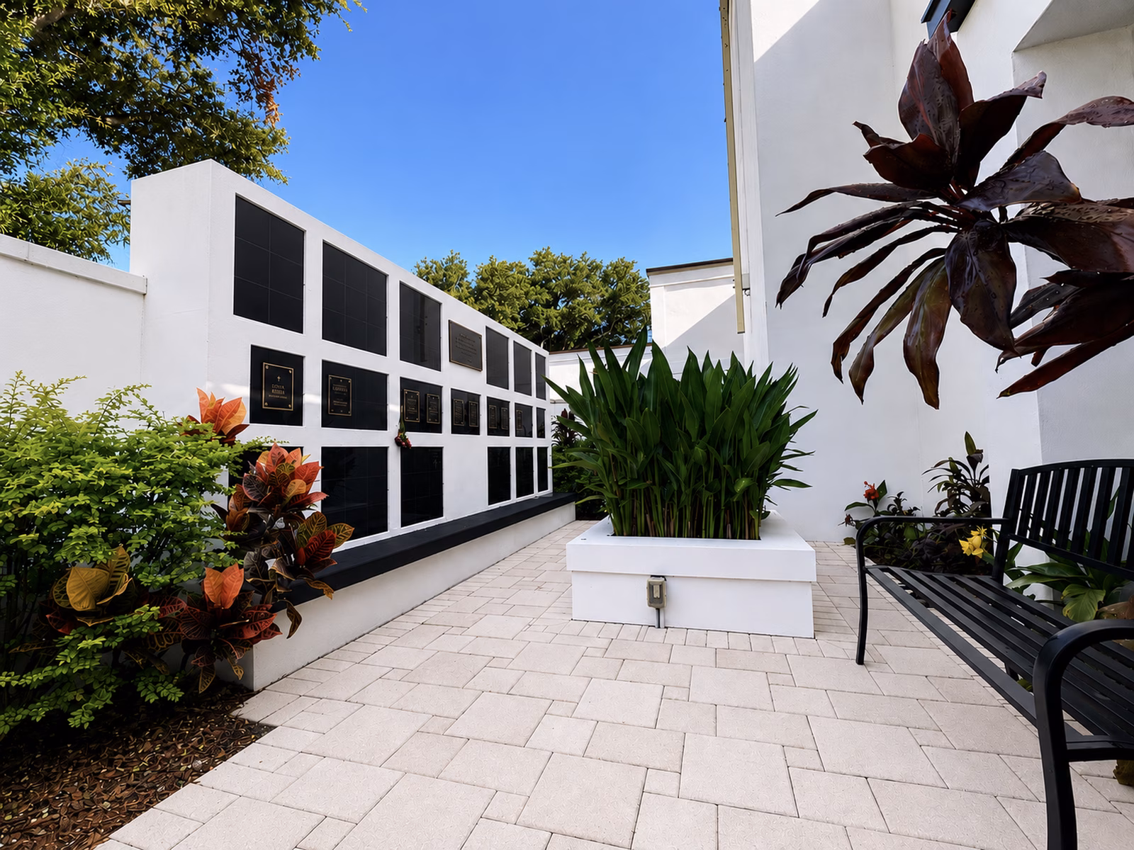 The Grace Lutheran columbarium: a low white outdoor wall set with rows of black granite niche faceplates, several engraved, bordered by croton and fern plantings along a paved courtyard.