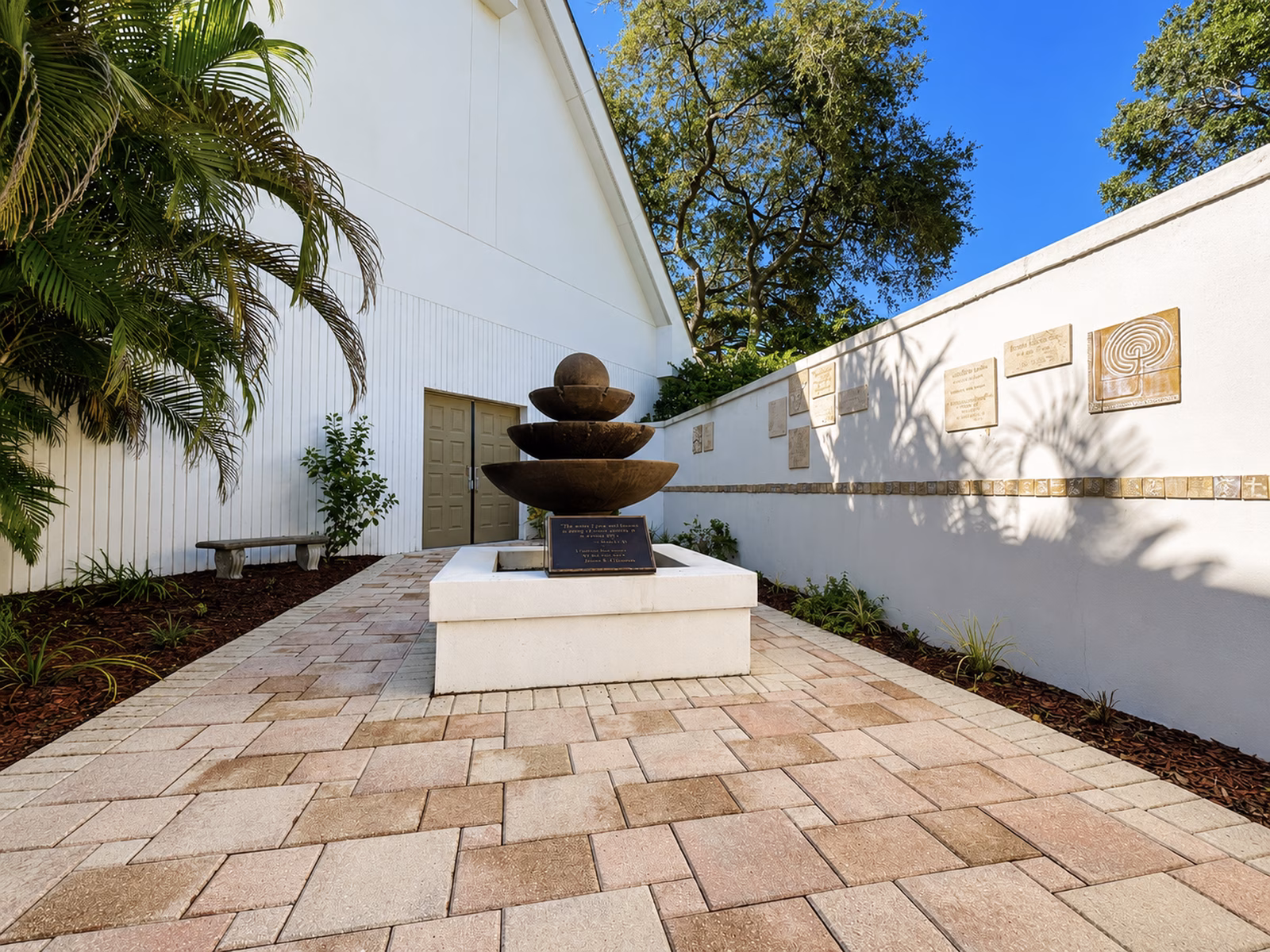 The Memorial Garden courtyard, with a three-tiered stone fountain on a low white base and engraved memorial plaques mounted in rows along the white garden wall.