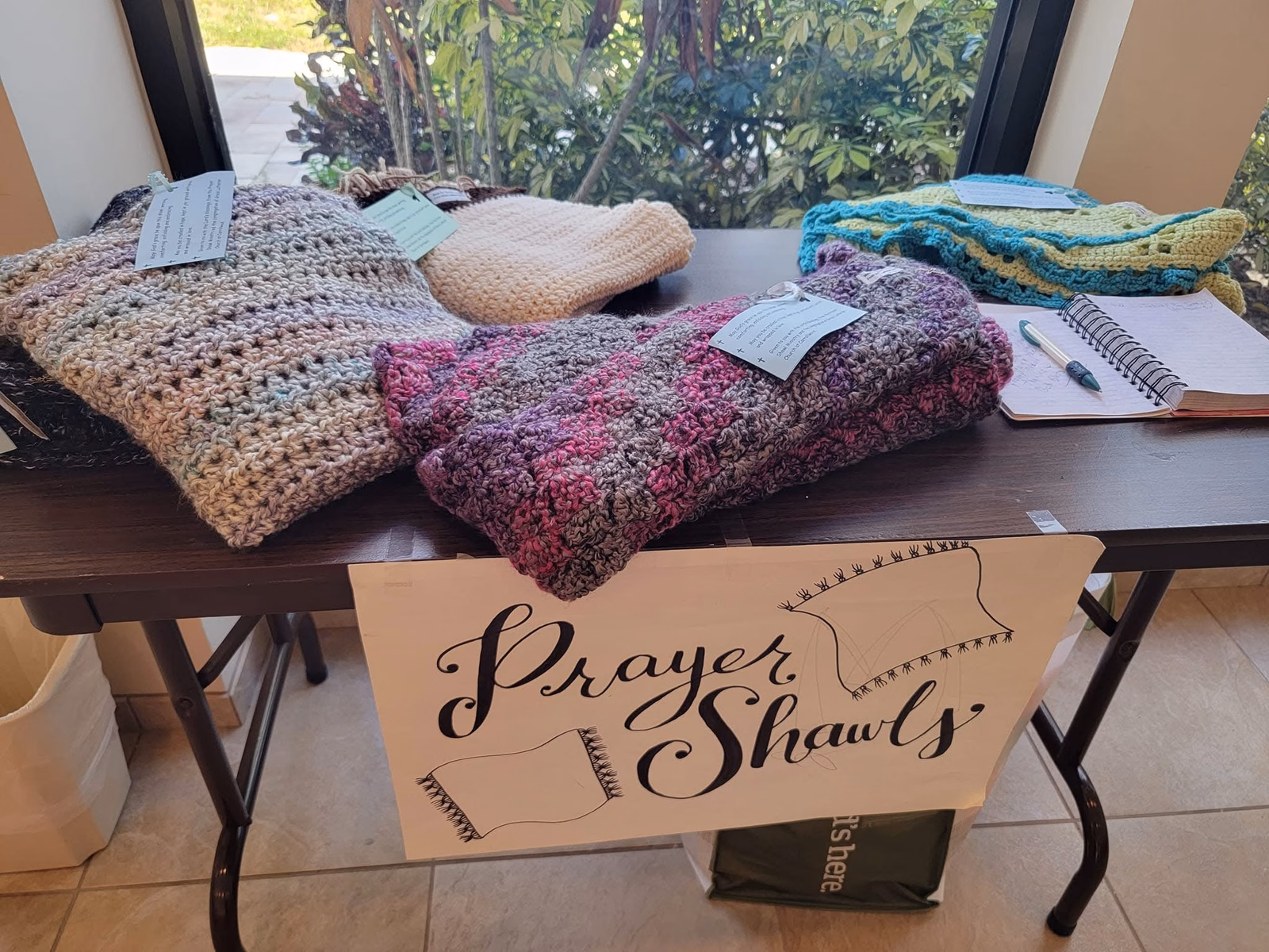 Handmade crocheted prayer shawls in pink, purple, cream, and teal folded on a table, each with a small note attached, behind a hand-lettered Prayer Shawls sign.