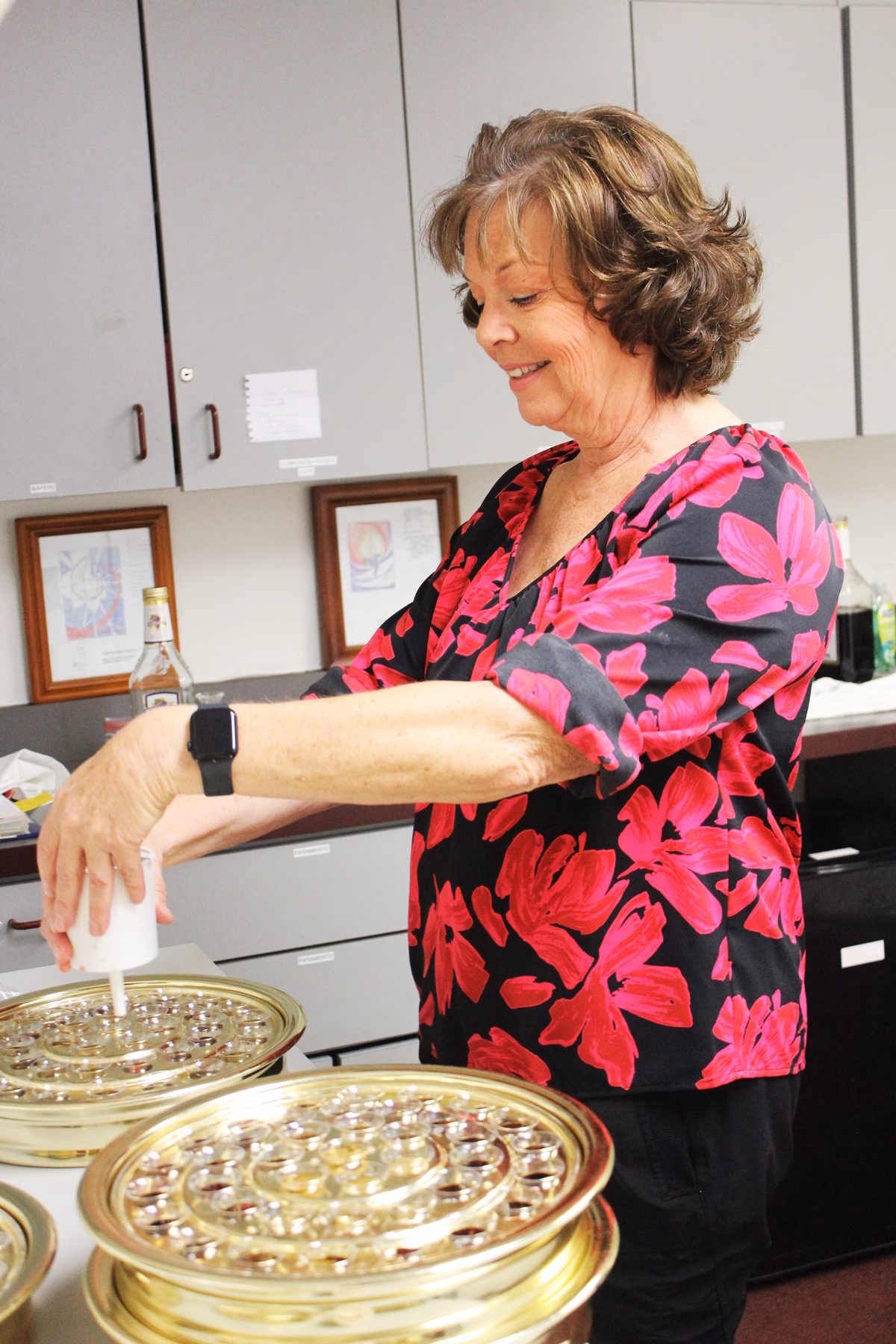 A smiling Altar Guild volunteer in a floral blouse filling individual communion cups in stacked brass communion trays, working at a counter in the church workroom.