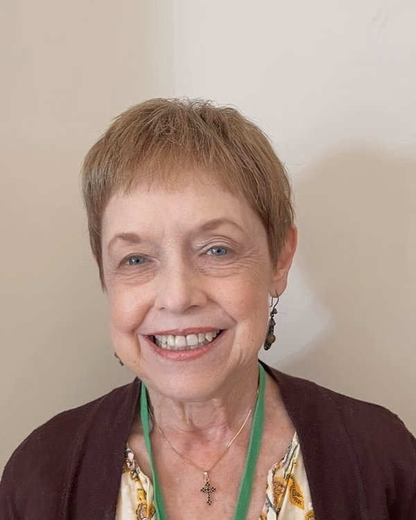 Barbara Campbell, smiling broadly, with short blonde hair, a dark burgundy cardigan and a small cross pendant.