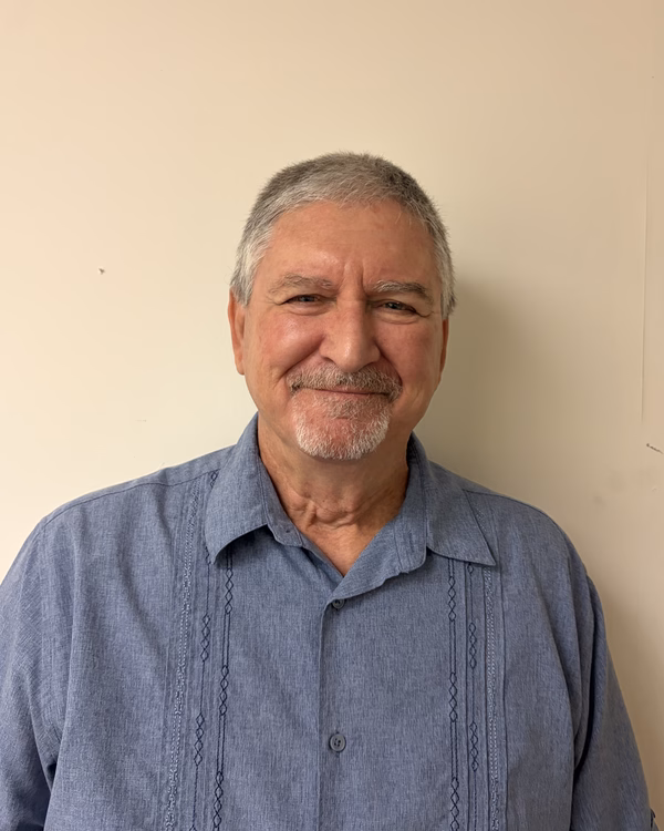 Brad Schmucker, smiling, with short grey hair and a mustache, wearing a blue pintucked short-sleeved shirt.