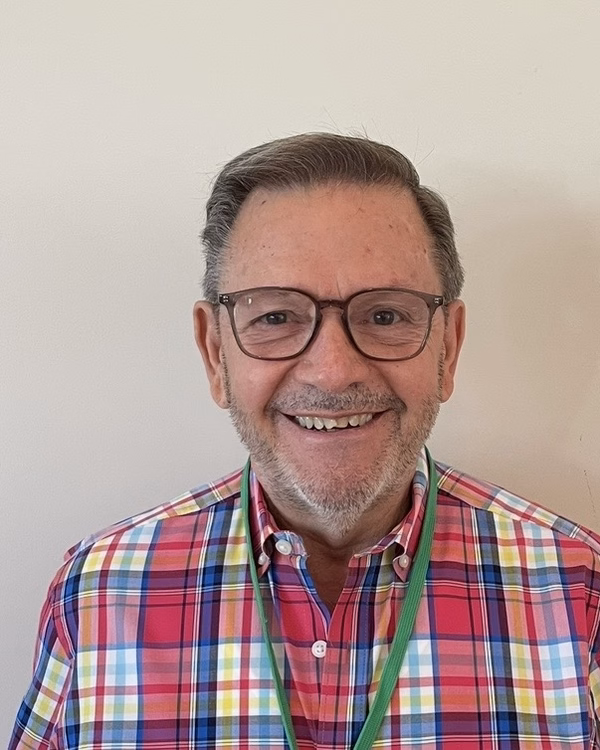 Juan Prat, smiling, with dark-framed glasses and a short grey beard, wearing a brightly colored plaid shirt.