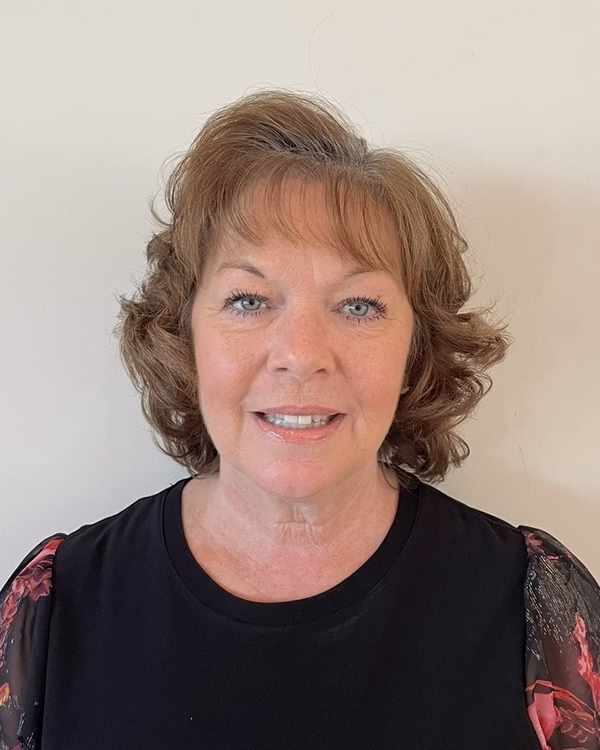 Liz Planz, smiling, with short curly auburn hair and a black top with floral sleeves.