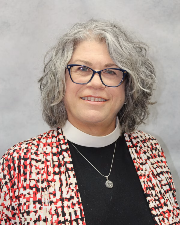 The Rev. Jennifer S. Michael, smiling, wearing a white clerical collar under a red, black and cream patterned jacket.