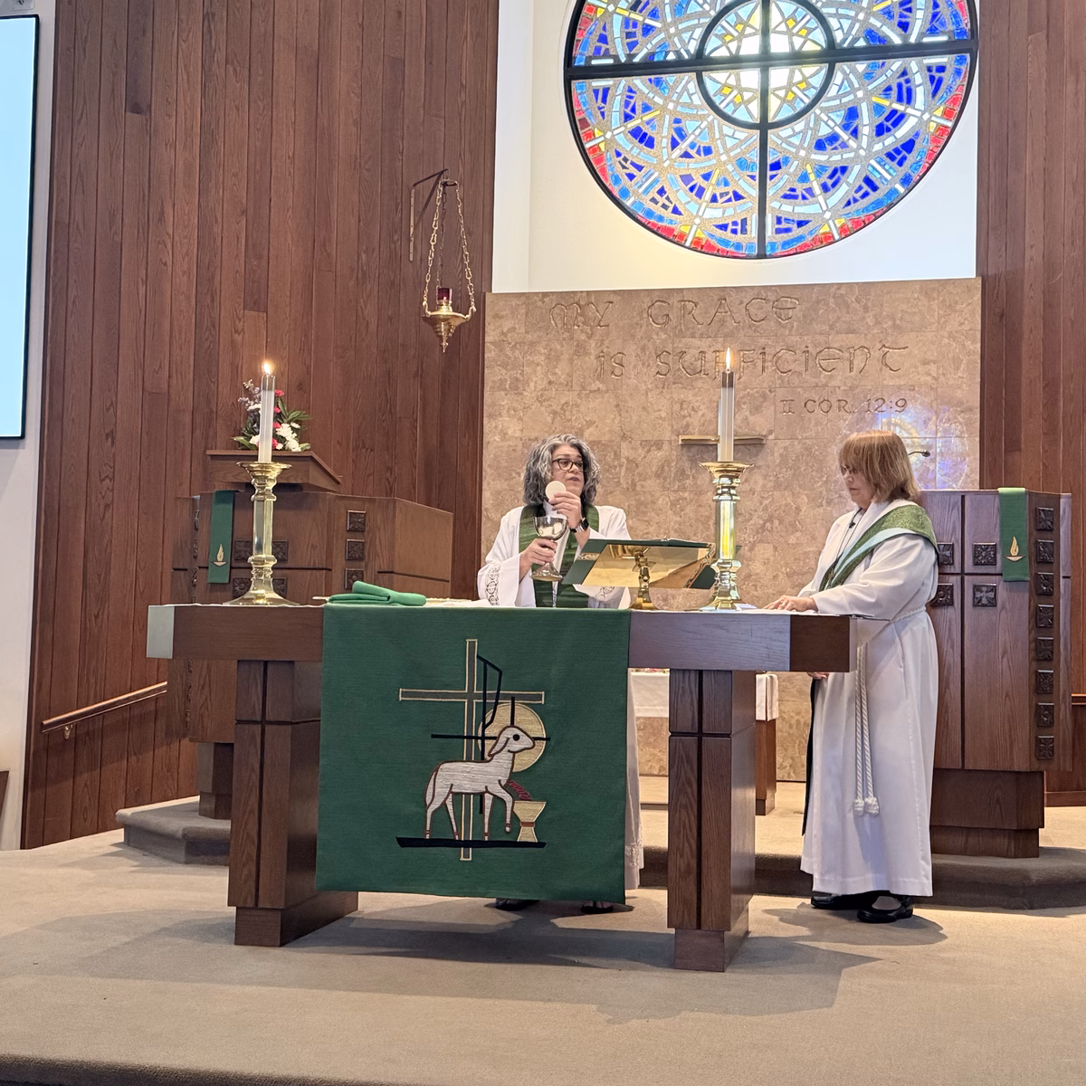 A pastor in a white alb and green stole raising the bread and chalice at the altar during communion, with an assisting minister in a white robe standing beside her beneath the round stained glass window and the words My Grace Is Sufficient.